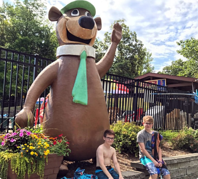 Two boys smile and pose beside a large bear statue at a vibrant water park