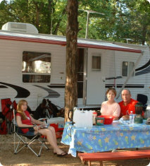 Three people sit at a picnic table and on chairs outside a camper trailer, with food and drinks on the table in a wooded campsite.