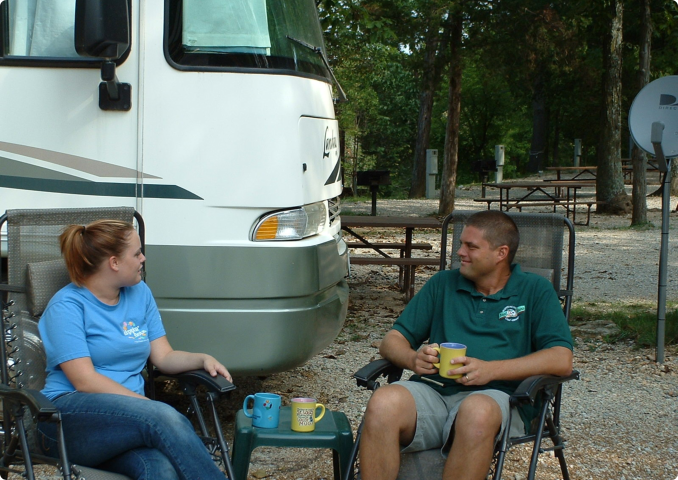 A man and woman sit in folding chairs by an RV, drinking from mugs. They appear relaxed, enjoying an outdoor camping setting with trees and picnic tables.
