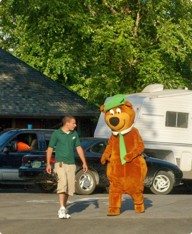 A man in a green hat and shirt walks alongside a bear mascot roasting marshmallows over a campfire
